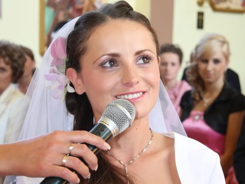 Panna młoda z welonem wypowiada przysięgę do mikrofonu, w tle goście w kościele